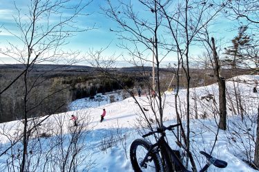 A snow-covered landscape with a black mountain bike parked in the foreground. In the background, skiers can be seen navigating a snowy slope under a clear blue sky. Leafless trees surround the scene, adding to the winter atmosphere. Batawa Trails mountain bike trail.