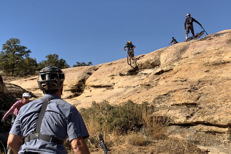 A group of mountain bikers navigating rocky terrain under a clear blue sky. Two riders are visible on a steep rock slope, while another is preparing to descend. In the foreground, a cyclist in a helmet and shirt is observing the scene. The landscape is characterized by rugged rocks and sparse vegetation, indicative of an outdoor adventure setting.