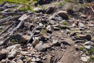 A rugged mountain bike path featuring scattered rocks and fallen logs, leading uphill. An orange mountain bike is parked on the side of the trail, with greenery visible amidst the rocky landscape and clear blue skies above. Ginny Trail mountain bike trail.