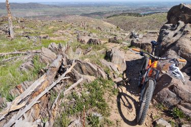 A mountain bike rests on a rocky trail surrounded by scattered tree stumps and boulders, with a panoramic view of green hills and valleys in the background under a clear blue sky. Ginny Trail mountain bike trail.