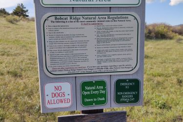 A sign at Bobcat Ridge Natural Area in Fort Collins, displaying regulations and guidelines for visitors. It includes information about park hours (open daily from dawn to dusk), activities that are prohibited such as bringing dogs and riding e-bikes, and contact information for emergencies. The sign is set against a background of open grassland with trees in the distance. Ginny Trail mountain bike trail.