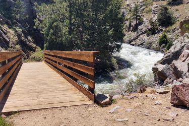 A wooden bridge extends over a river, surrounded by rocky terrain and tall green trees under a blue sky with fluffy clouds. The rushing water can be seen flowing beneath the bridge, creating a serene natural setting. Walker Ranch mountain bike trail.