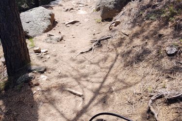 A view from above a mountain bike on a narrow, sandy trail surrounded by rocks and trees. The bike's handlebars and front wheel are visible in the foreground, while the path ahead winds through a natural landscape. Soft sunlight filters through the trees, creating dappled shadows on the ground. Walker Ranch mountain bike trail.
