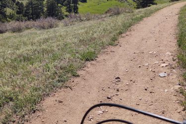 A view from the handlebars of a mountain bike on a dirt trail, surrounded by green hills and pine trees under a partly cloudy sky. Walker Ranch mountain bike trail.