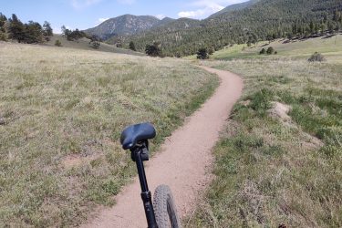 A mountain biking view showing a dirt trail winding through a grassy meadow, with mountains in the background under a blue sky with scattered clouds. The image captures the seat and post of a bike in the foreground, emphasizing a sense of adventure in nature. Walker Ranch mountain bike trail.
