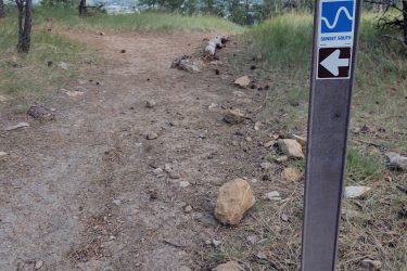 A dirt trail surrounded by pine trees, with a trail marker indicating the path number 014 and the direction "Sunset South." The scene includes rocky terrain and a clear sky. HLMP mountain bike trail.