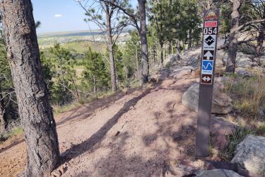 A hiking trail marker situated along a dirt path in a forested area, surrounded by tall pine trees. The sign features various trail symbols, including markers for different routes, with a scenic view of hills and a valley in the background under a clear blue sky. HLMP mountain bike trail.