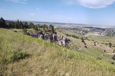 A scenic view from a hillside overlooking a valley, with a mix of green grass, scattered trees, and rocky cliffs. The distant landscape features a town and open fields under a bright blue sky with a few clouds. HLMP mountain bike trail.
