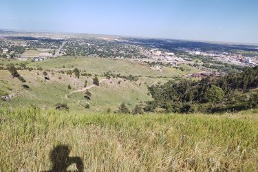 A panoramic view from a hillside looking down on a sprawling city landscape with green hills and patches of trees in the foreground. A person's shadow is visible in the lower corner as they hold a camera, capturing the scenic vista. HLMP mountain bike trail.