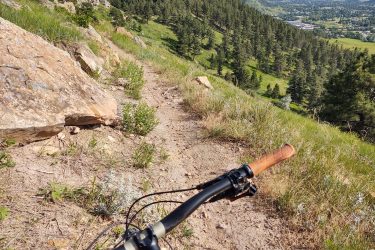 A close-up view of a mountain bike handlebar with a wooden grip, positioned on a narrow dirt trail surrounded by rocky terrain and lush green vegetation, overlooking a scenic valley with trees and distant hills under a clear blue sky. HLMP mountain bike trail.
