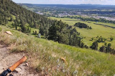 A scenic view of a lush, green hillside with pine trees, overlooking a valley and distant mountains. A portion of dirt trail is visible in the foreground, along with the grip of a bike handlebar. The sky is partly cloudy, enhancing the natural beauty of the landscape. HLMP mountain bike trail.