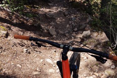 A mountain bike with an orange frame is positioned on a rocky, unpaved trail surrounded by trees and underbrush, providing a view looking down the trail ahead. Sunlight filters through the foliage, illuminating the terrain. Lion Gulch mountain bike trail.