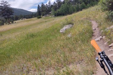 A scenic view of a grassy trail winding through a landscape with scattered rocks, surrounded by trees and mountains under a partly cloudy sky. The image captures the perspective of a bicycle handlebar in the foreground, suggesting an outdoor cycling adventure. Lion Gulch mountain bike trail.