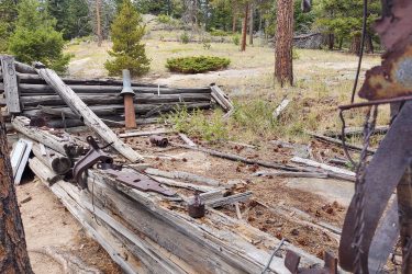 Abandoned wooden structure in a forested area, surrounded by trees and scattered debris, including rusted metal objects and fallen logs. Pine cones are visible on the ground among the natural vegetation. Lion Gulch mountain bike trail.