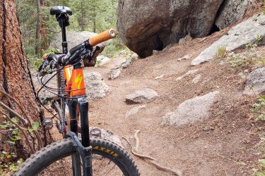 A mountain bike rests against a tree along a narrow dirt path surrounded by tall green trees and large boulders. The trail appears rugged with rocks and roots scattered throughout, indicating a natural outdoor setting ideal for biking. Lion Gulch mountain bike trail.