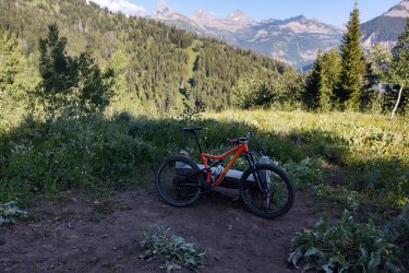 A mountain bike resting on a wooden bench in a lush, green landscape, with rolling hills and mountain peaks in the background under a clear blue sky. The scene captures the beauty of nature, ideal for outdoor activities. Grand Targhee Bike Park mountain bike trail.