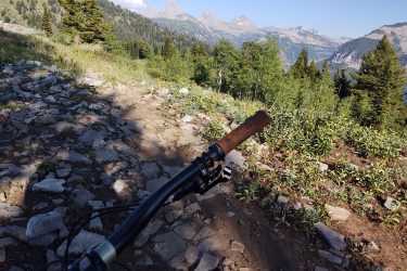 A view from the handlebars of a mountain bike on a rocky trail, with a scenic backdrop of mountains and lush greenery under a clear blue sky. Grand Targhee Bike Park mountain bike trail.