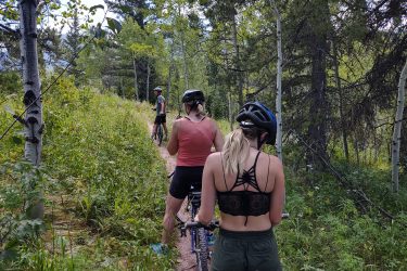 Three cyclists pause along a forest trail surrounded by greenery and trees. Two women are seen from behind, wearing helmets and casual athletic clothing, while a third cyclist is further ahead. The path is narrow and lined with plants, suggesting a serene outdoor environment. Rush Hour mountain bike trail.