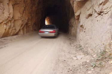 A car driving through a dirt road tunnel carved into rocky terrain, with rough stone walls and a light visible at the end of the tunnel. Captain Jack's mountain bike trail.