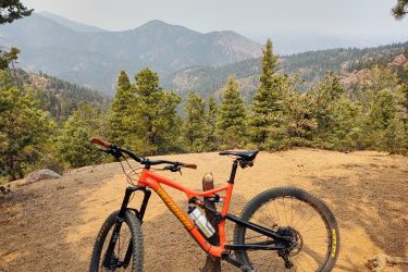 A mountain bike resting on a dirt trail with a panoramic view of misty mountains and pine trees in the background. The bike has an orange frame and is positioned at the edge of a scenic overlook. Captain Jack's mountain bike trail.