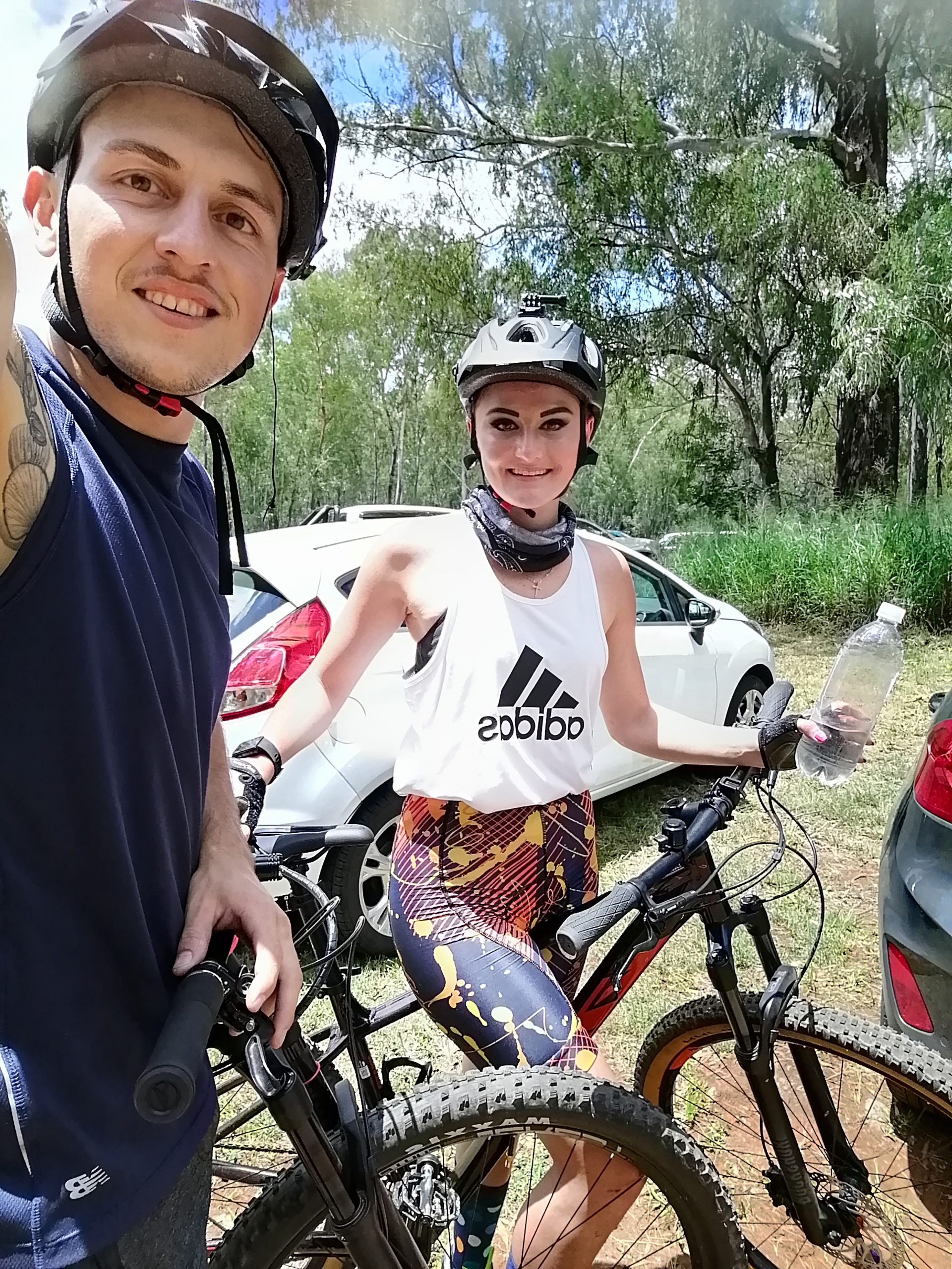 A young man and woman pose together for a selfie, both wearing helmets and athletic clothing. They are standing next to their mountain bikes in a natural outdoor setting, with trees and parked cars in the background. The woman holds a water bottle while smiling at the camera. Red Barn mountain bike trail.