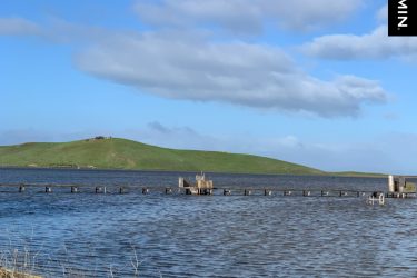 A scenic view of a calm body of water with a dock extending into it. In the background, there is a lush green hill under a partly cloudy blue sky. The foreground features rocks along the shoreline and a few structures at the end of the dock. Coyote Hills Regional Park mountain bike trail.