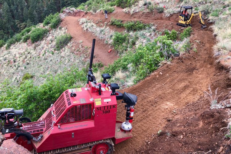 A red trail building machine is positioned on a hillside, with a worker operating a small excavator in the background. The machine is designed to help shape and maintain dirt paths, and the area shows freshly cleared soil amidst greenery and rocky terrain.