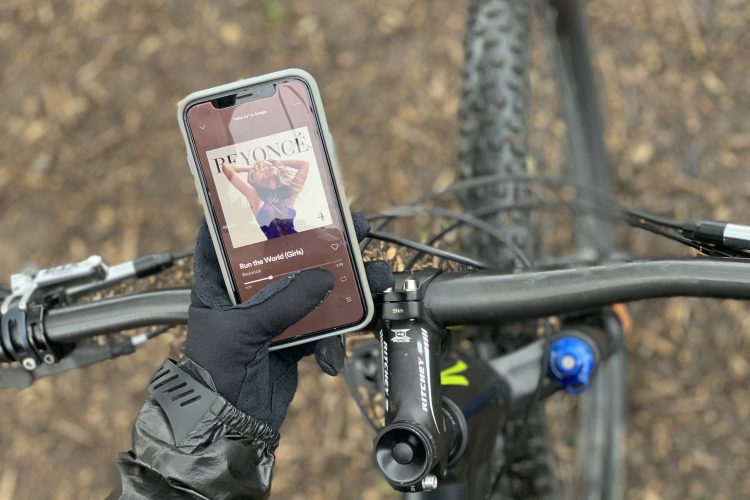 A close-up view of a person's hand wearing a black glove holding a smartphone displaying the cover of the song "Run the World (Girls)" by Beyoncé. The phone is positioned on the handlebars of a bicycle, with a blurred background of a dirt path and bike components.