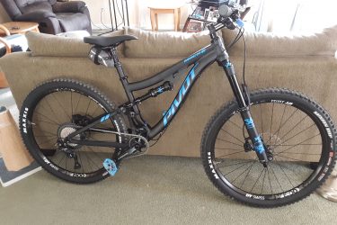 Pivot Mach 6: A black mountain bike with blue accents stands beside a couch in a living room. The bike features wide tires, a sturdy frame, and a front suspension fork, indicating it's designed for off-road use. In the background, a cozy seating area is visible, along with a coffee table and various household items.