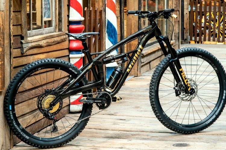 A black mountain bike with gold accents is leaning against a wooden wall. The bike features thick, textured tires designed for off-road riding, a suspension system, and a water bottle holder. The background includes wooden structures and a barbershop pole, suggesting a rustic or outdoor setting.