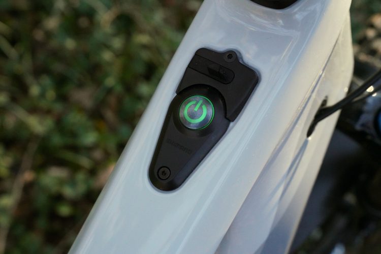 Close-up of a power button on an electric bike, featuring a green illuminated icon, set against a blurred natural background with greenery.