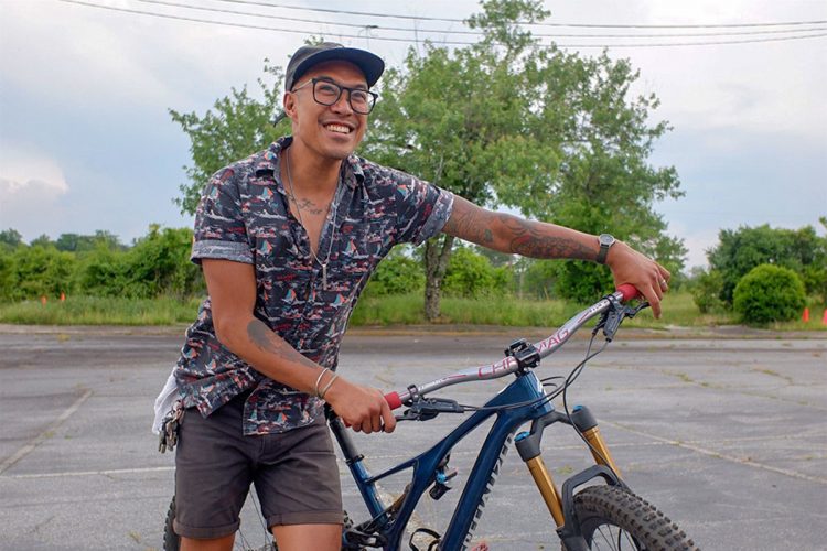 A person standing next to a mountain bike in an outdoor setting, smiling and wearing a short-sleeved, patterned shirt and shorts. They have tattoos visible on their arms and are wearing glasses and a cap. The background features greenery and an open space with a slightly overcast sky.