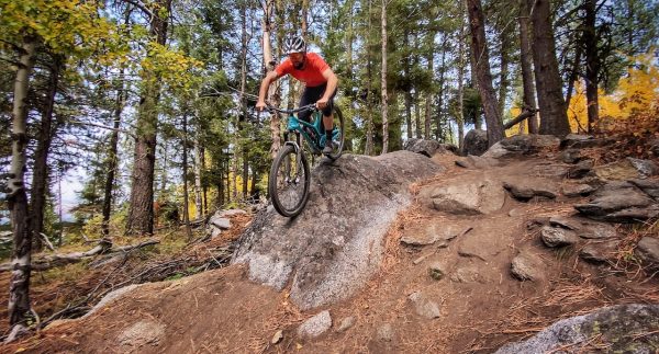 A mountain biker in a red shirt and helmet navigates a rocky trail in a forested area, airborne over a large boulder with trees and autumn foliage in the background.