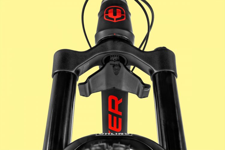 A close-up view of a bicycle fork, showcasing the black suspension arms and a red logo on the fork's steerer tube. The background is a solid light yellow color.