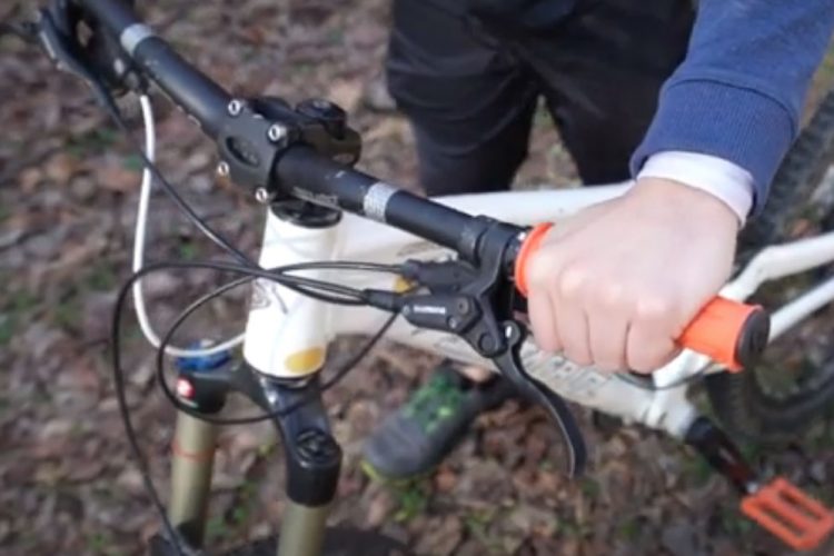 A close-up view of a person's hand gripping the handlebar of a mountain bike, showcasing the brake lever and orange grip. The background features a forest floor with fallen leaves.