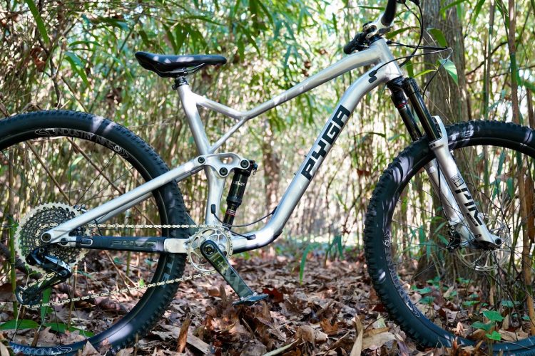 A silver mountain bike with a sleek frame and wide tires, resting on a bed of colorful leaves in a dense forest. The bike features detailed components, including a suspension system and a prominent brand logo. Surrounding vegetation adds a natural background to the scene.