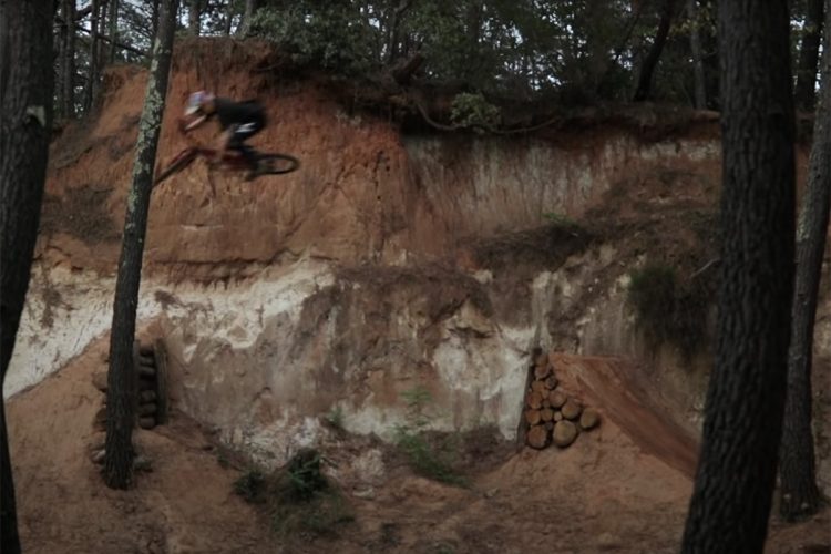 A mountain biker performing a jump over a dirt ramp in a wooded area, with steep earthen slopes and trees in the background.