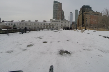 A snowy urban landscape featuring a path covered in snow with a view of modern skyscrapers. In the background, a historic building with large windows and a clock tower is visible, along with tall buildings like One World Trade Center. The scene is set under a gray sky, conveying a chilly winter atmosphere. West Street Greenway mountain bike trail.