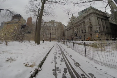 A snow-covered path winds through a park in an urban setting, flanked by bare trees and tall buildings. Footprints and tire tracks are visible in the fresh snow, with a fence along one side. The scene captures a quiet winter day, with a gray sky overhead. West Street Greenway mountain bike trail.