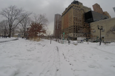 A snowy urban landscape featuring a path winding through a park, surrounded by bare trees and buildings. The scene is set under a cloudy sky, with snow covering the ground and creating a tranquil winter atmosphere. West Street Greenway mountain bike trail.