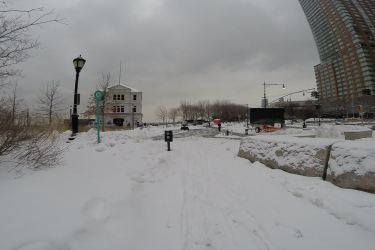A snow-covered pathway leading towards a historic building, with snow-laden trees and a streetlamp on the left. In the distance, a modern building rises against a cloudy sky. Footprints are visible in the fresh snow, and a traffic signal can be seen in the background, indicating a winter scene in an urban environment. West Street Greenway mountain bike trail.