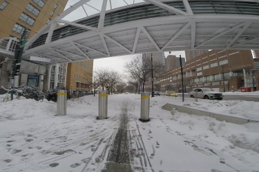 A snow-covered urban scene showing a pathway lined with snow and tire tracks, flanked by concrete barriers and trees. Above, a modern pedestrian bridge is visible. In the background, buildings and parked cars are partially obscured by snow, with a hint of a cloudy sky. West Street Greenway mountain bike trail.
