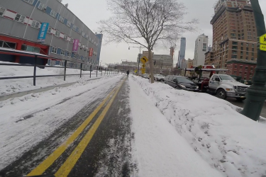 A snowy urban street featuring a bike lane bordered by snow piles on either side. In the background, a mix of low-rise and high-rise buildings can be seen, with some displaying colorful banners. A person is walking along the sidewalk, while parked and moving vehicles are visible to the right. The sky appears overcast, contributing to the winter atmosphere. West Street Greenway mountain bike trail.