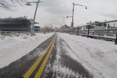 A snow-covered bike path lined with snowbanks, leading towards a ship in the background. The scene is set on a cloudy day with a mixture of urban structures and greenery visible along the path, highlighting a winter landscape. West Street Greenway mountain bike trail.