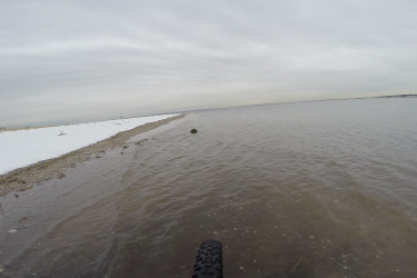 A calm beach scene showing a narrow strip of sand and water with a snowy area in the foreground. The landscape is overcast, with gray skies reflecting on the surface of the water, creating a serene atmosphere. A bicycle tire is visible in the bottom corner of the image, suggesting an outdoor activity such as biking along the waterfront. Gateway Beach mountain bike trail.