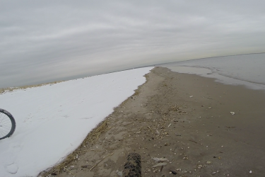 A bicycle is positioned on the left side of the image, alongside a shoreline that transitions from snow to sandy beach. The scene features a cloudy sky and calm water. The shore appears sparsely dotted with shells and debris. Gateway Beach mountain bike trail.
