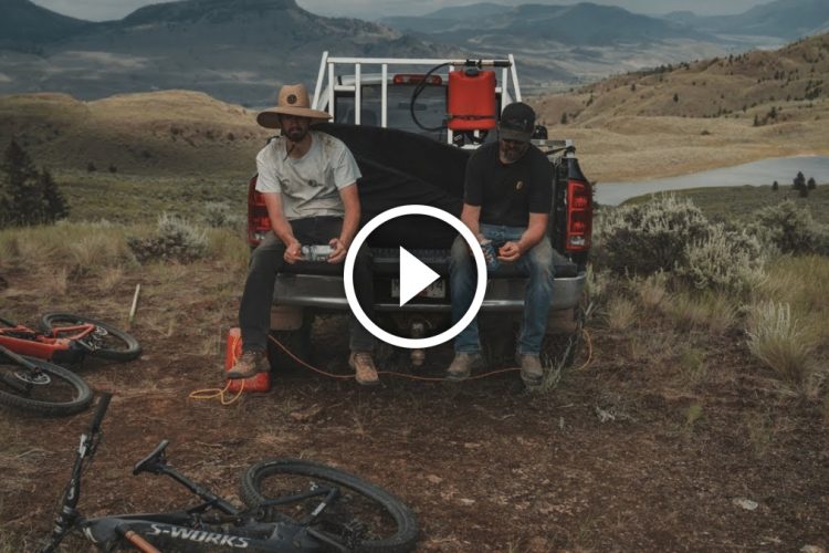 Two men sit on the back of a pickup truck on a hillside, enjoying a scenic view of mountains and a lake. They are casually dressed, with one wearing a wide-brimmed hat. Mountain bikes are resting on the ground nearby. The scene conveys a sense of relaxation and outdoor adventure.