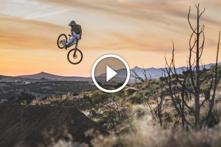 A mountain biker performing an aerial stunt over a dirt jump at sunset, with hills and a colorful sky in the background.