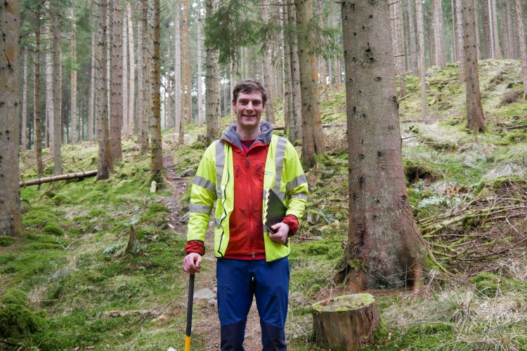 A person standing on a forest trail surrounded by tall trees, wearing a bright yellow safety jacket over a red shirt, blue pants, and sturdy boots. They are holding a clipboard in one hand and a tool in the other, smiling at the camera. The forest floor is covered in moss and fallen leaves, creating a natural setting.