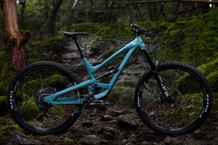 A mountain bike with a teal frame is positioned on rocky terrain, surrounded by lush moss and trees. The bike features wide tires and suspension components, showcasing its rugged design suitable for off-road trails. Natural light filters through the forest, highlighting the bike's details against the earthy background.